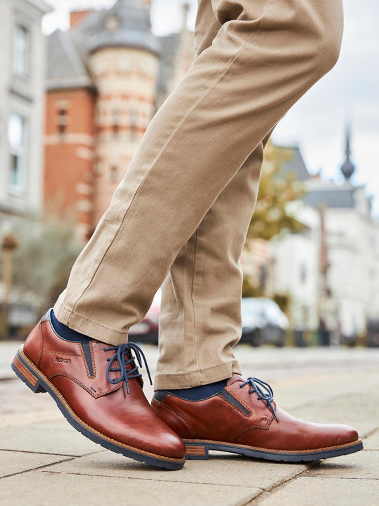 En person i brune bukser og brun 02142 Rieker Leather Amaretto Dressko by Rieker Comfort & Style, med blå lisser og vid passform, går langs en bygate med historiske bygninger.