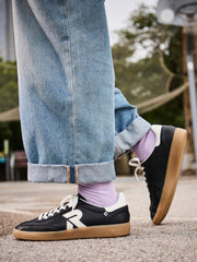 En person står utendørs på en basketballbane iført løse blå jeans med mansjetter, lyselilla sokker og Rieker Sport 03470 San Diego skinnsko til dame, svart med hvite detaljer og gummisåler.