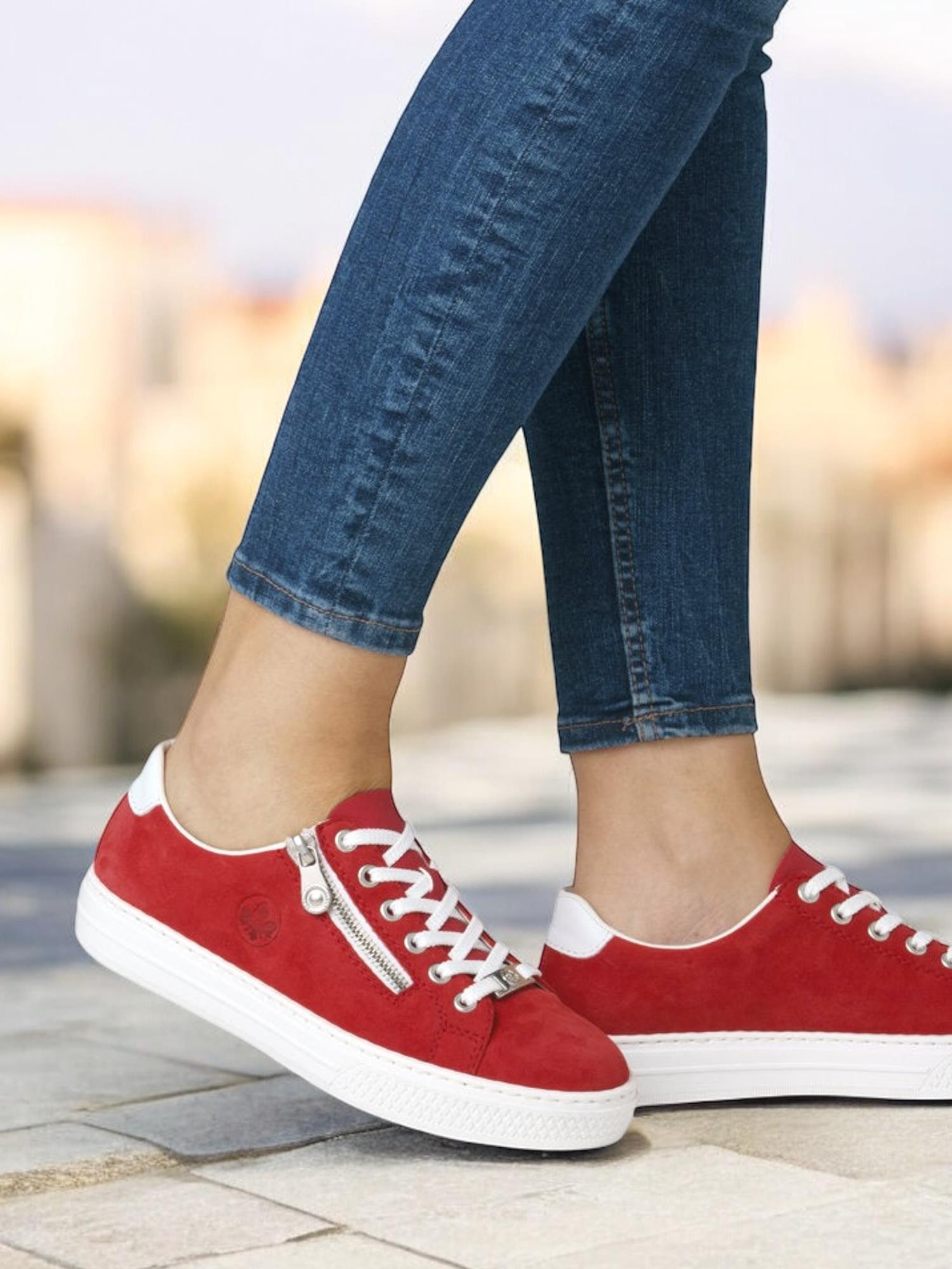 En person står utendørs på fliser, iført blå skinny jeans og Rieker 03155 Red Leather Memory Sneakers med hvite såler og lisser og en behagelig MemoSoft-innersåle.