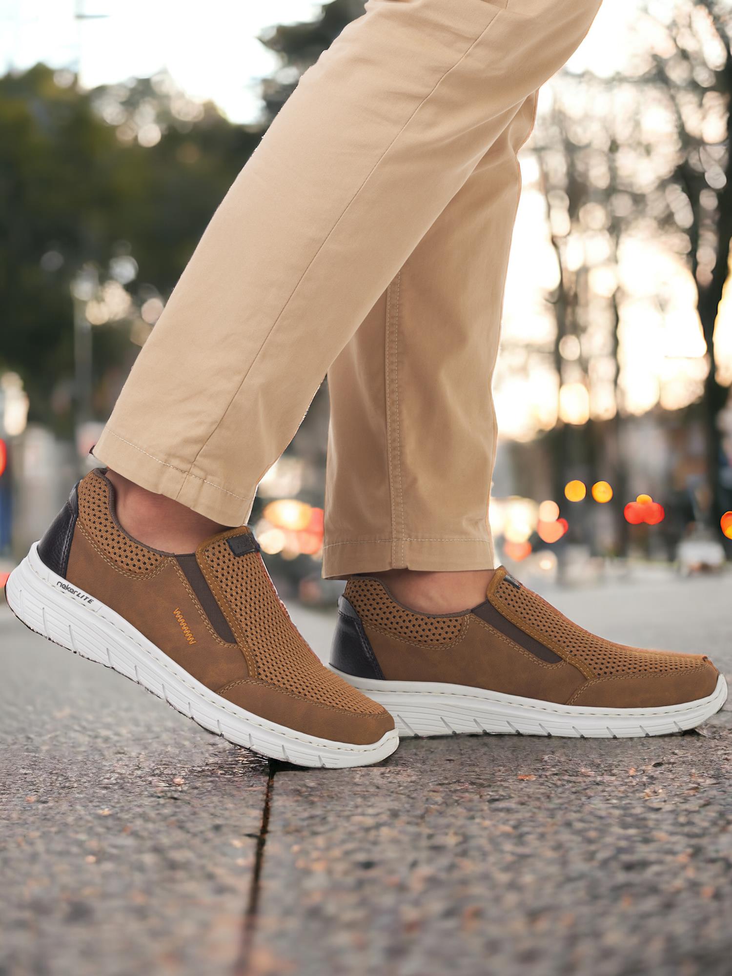 Noen som har på seg Rieker Comfort & Style 07529 brune slip-on antistress-sneakers med beige bukser går utendørs på en bygate i skumringen, med uskarpe lys og trær i bakgrunnen.