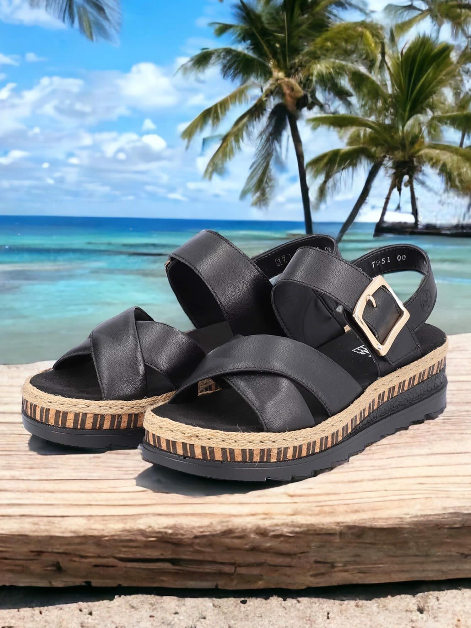 08084 Rieker Arizona Sandal Black Leather fra Rieker Comfort & Style, med tykke såler og spenneremmer, byr på ekstra myk komfort mens den hviler på tre med en tropisk strand, blått hav og palmer i bakgrunnen.