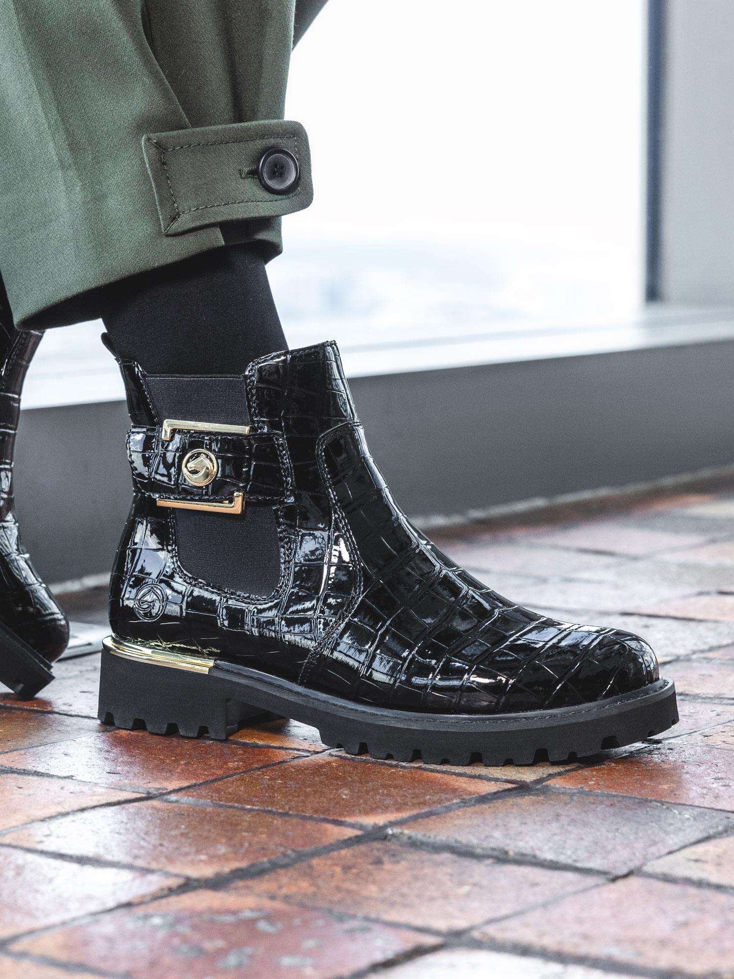 En person har på seg 05024 Remonte Gold Croc Boot fra Remonte, med lakkskinn og svart krokodillemønster med en liten lomme og gulldetaljer, sammen med en grønn bukse på et flislagt gulv.