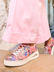 En person i lyserosa vide bukser og dristige 03284 Rieker ColorSplash-sneakers fra Rieker Comfort & Style, med et fargerikt design, hvite lisser og en glidelås på siden, står på en steintrapp.