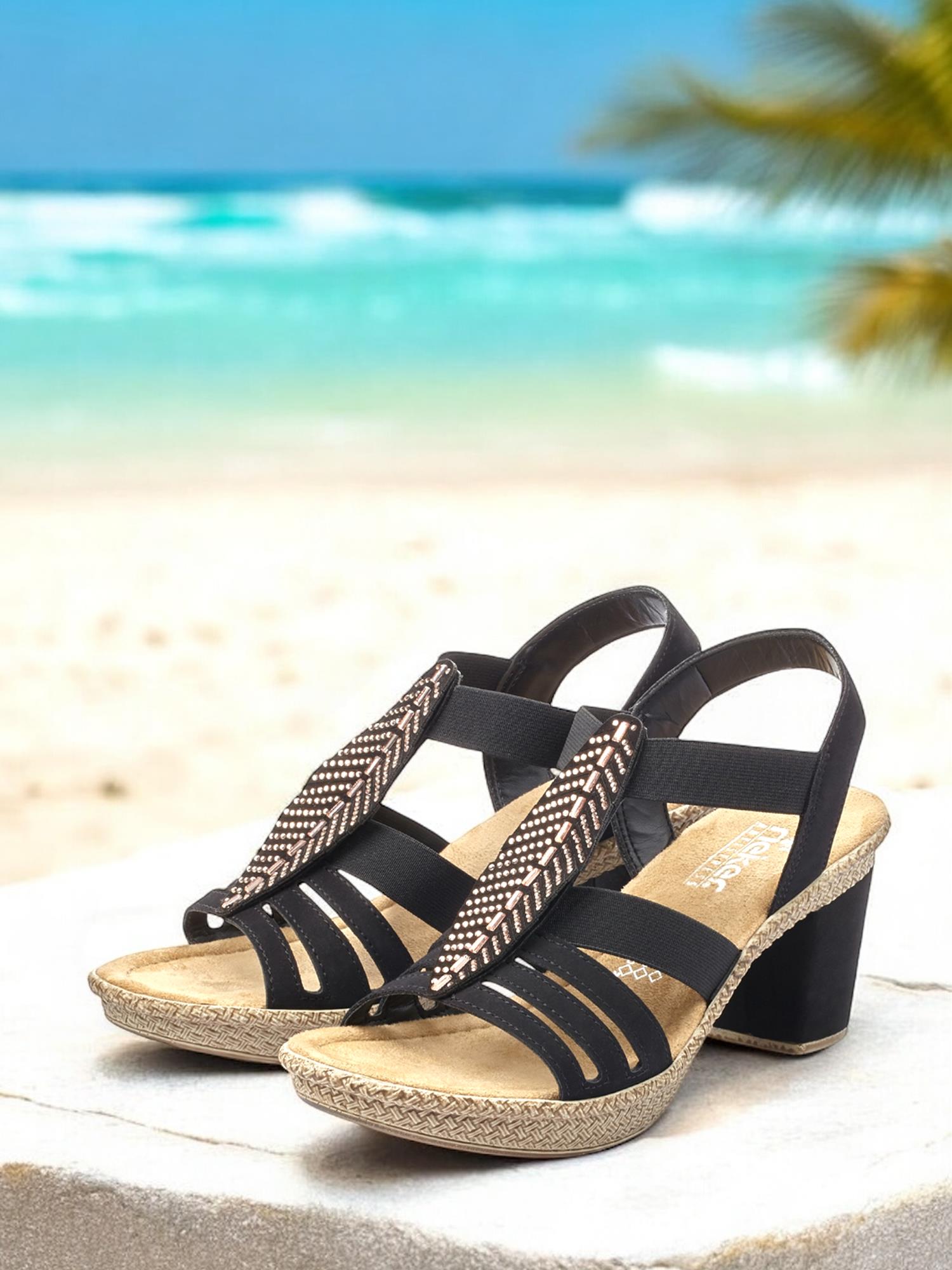07498 Rieker Feather Heeled Sandal fra Rieker Comfort & Style, med mønstrede stropper og komfort, står på en steinavsats med en sandstrand, turkise bølger og et palmetre i bakgrunnen.