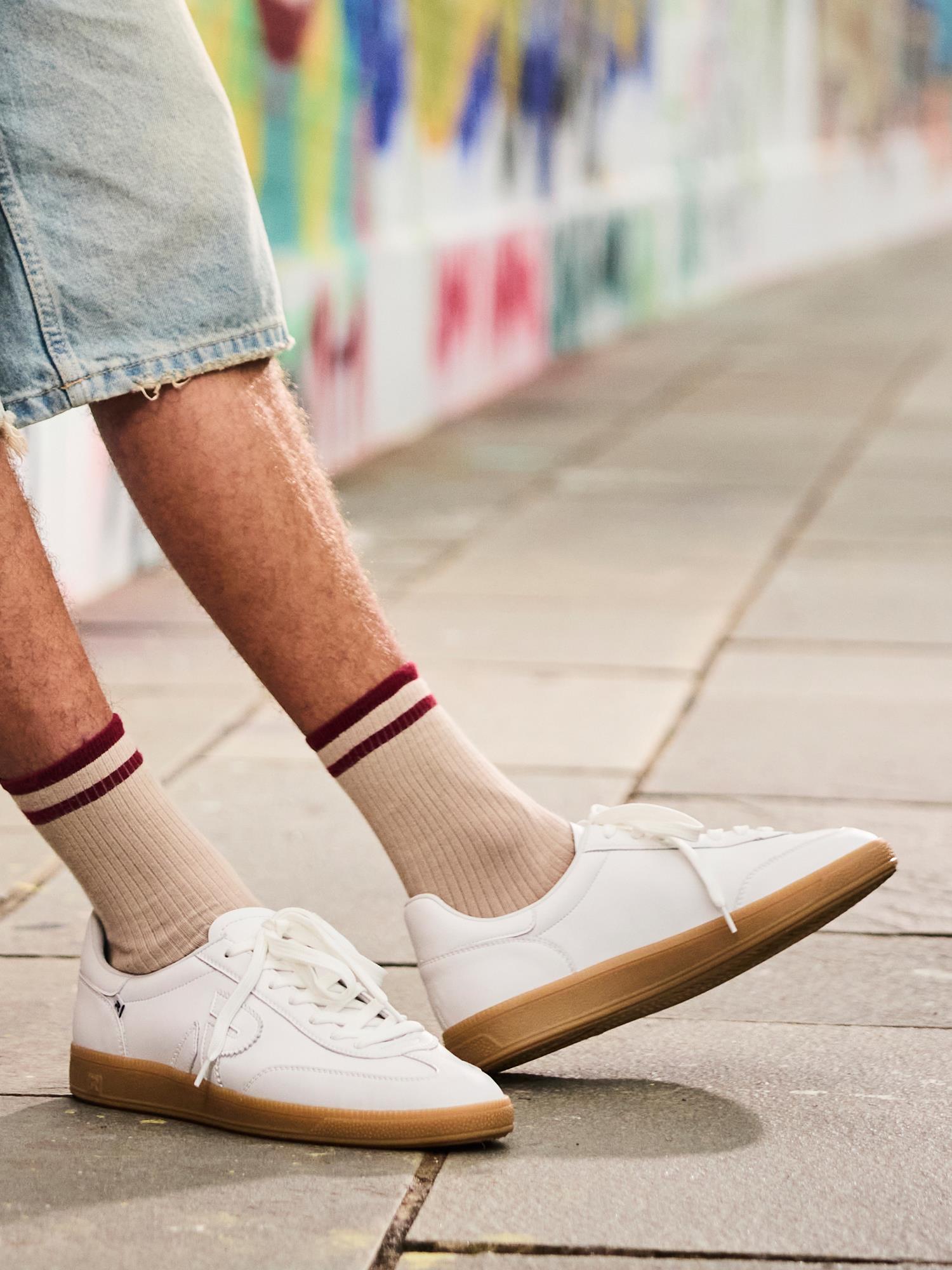 En person i beige sokker med røde striper, denimshorts og hvite Rieker 03336 San Diego Leather Sneakers for men går på et asfaltert fortau ved siden av en fargerik graffitikledd vegg.