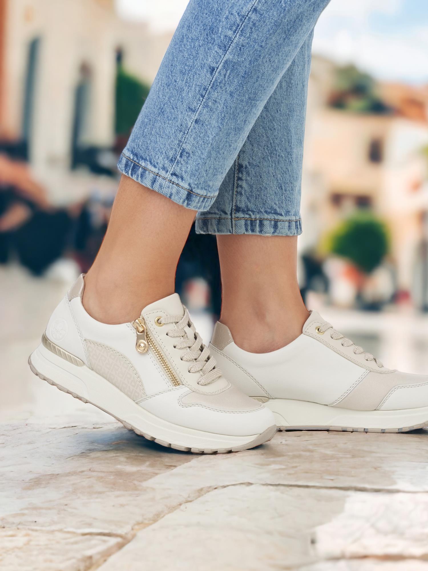 En person iført lyse 07583 Rieker Reflect Leather Zip Sneakers by Rieker Comfort & Style og blå jeans står utendørs på en steinflate, med bygninger og grøntområder uskarpt i bakgrunnen.