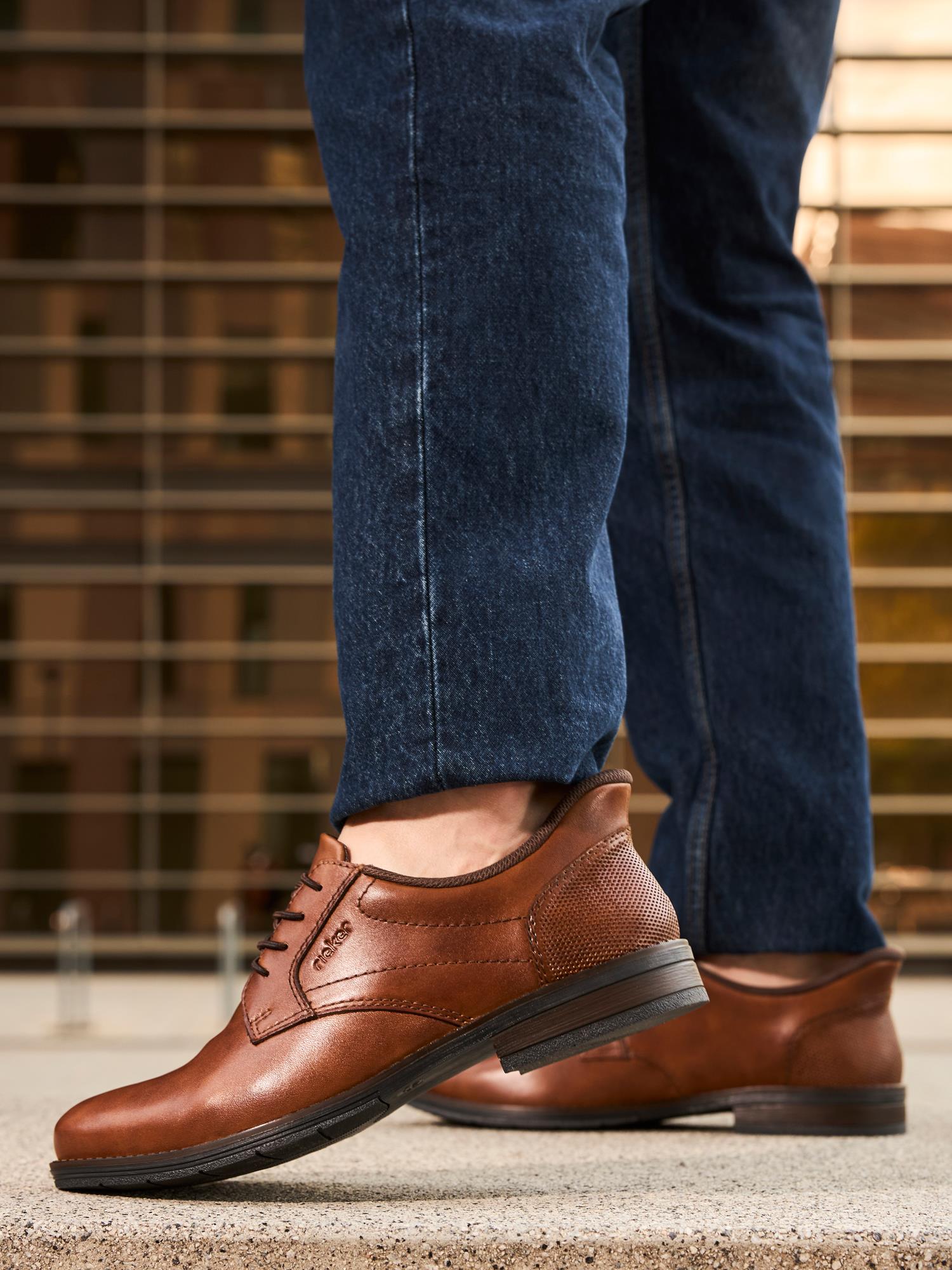 En person i mørkeblå jeans og brune 02889 Rieker EasyWalk Slip-Ons fra Rieker Comfort & Style står på betong, med en uskarp bygning og metallrekkverk i bakgrunnen.