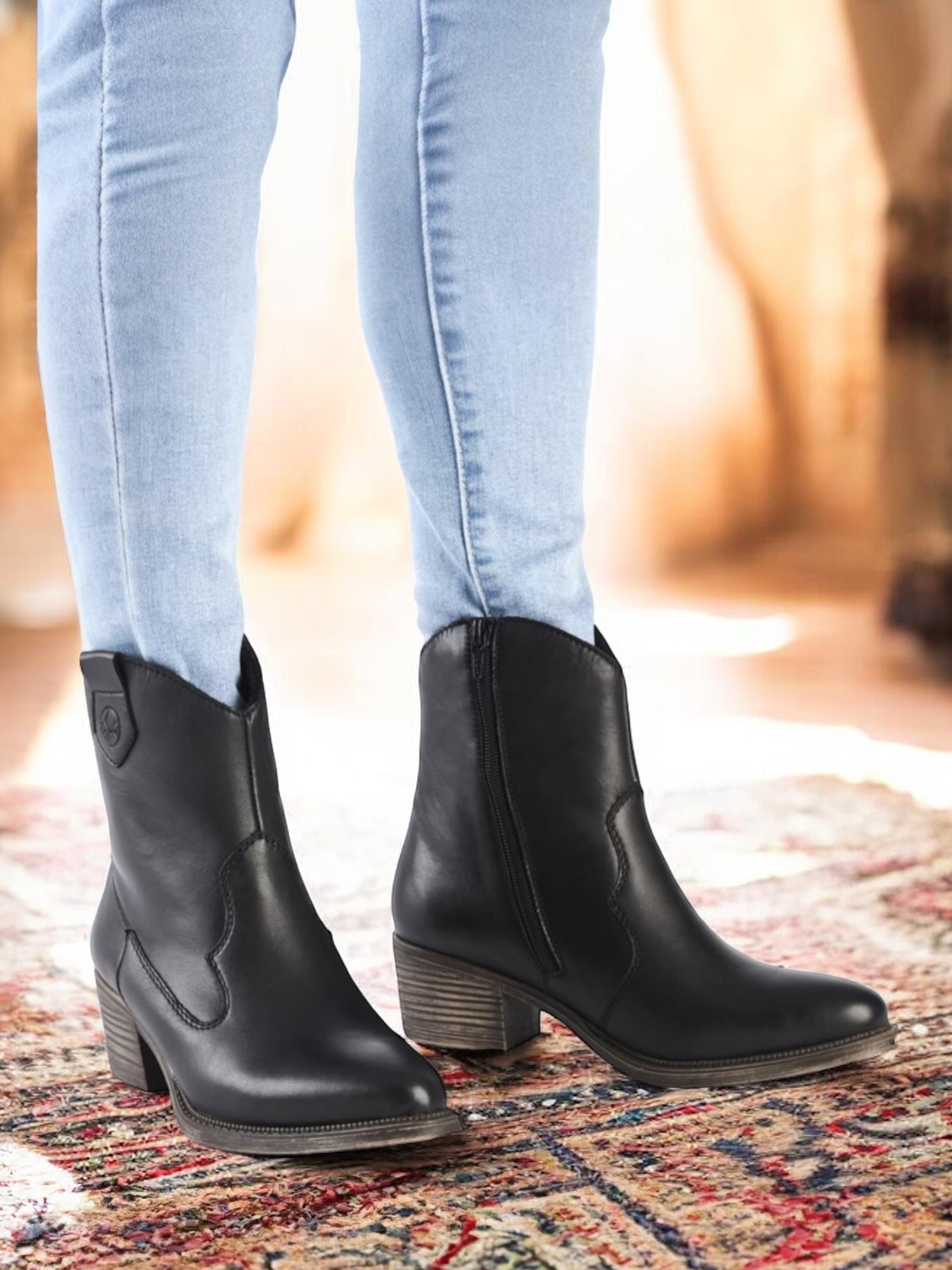 En person i lyseblå skinny jeans og 04859 Rieker Leather Cowgirl Boots fra Rieker Comfort & Style med lav blokkhæl står på et mønstret teppe i et sterkt opplyst rom.